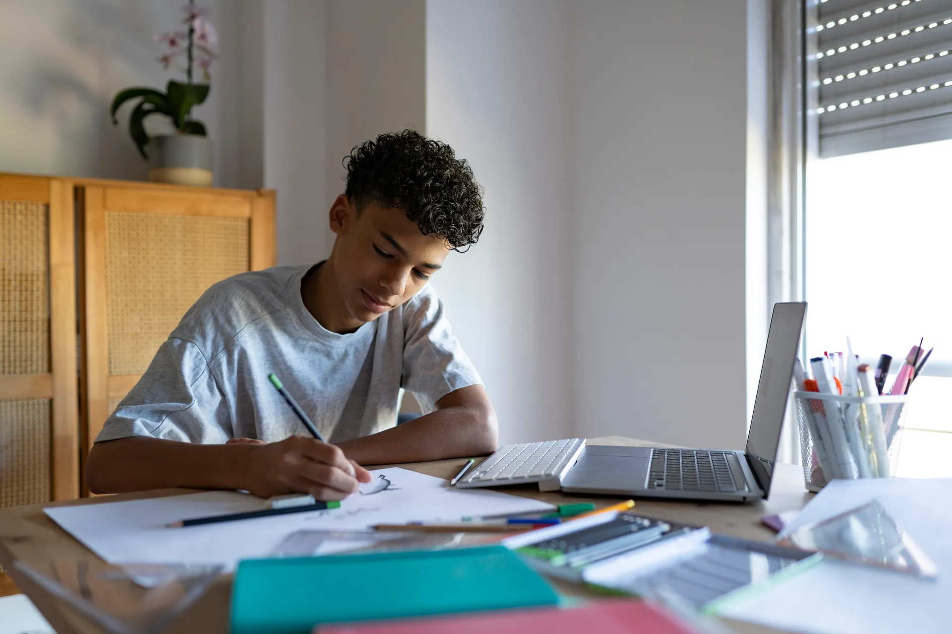 A teenage student in uniform studying on a laptop at a sunlit desk at home