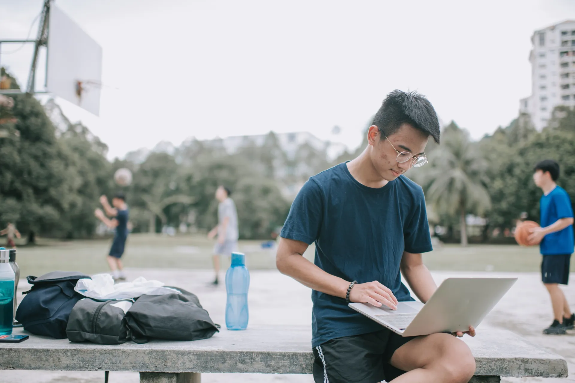 A Vaughan College student doing online coursework on a laptop outdoors while friends play basketball nearby