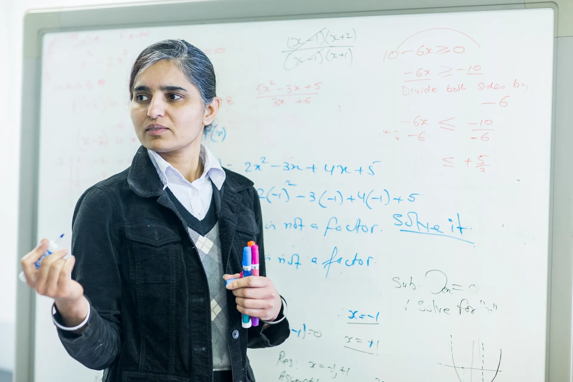 A Vaughan College math teacher mid-explanation, holding markers, equations on the whiteboard behind her