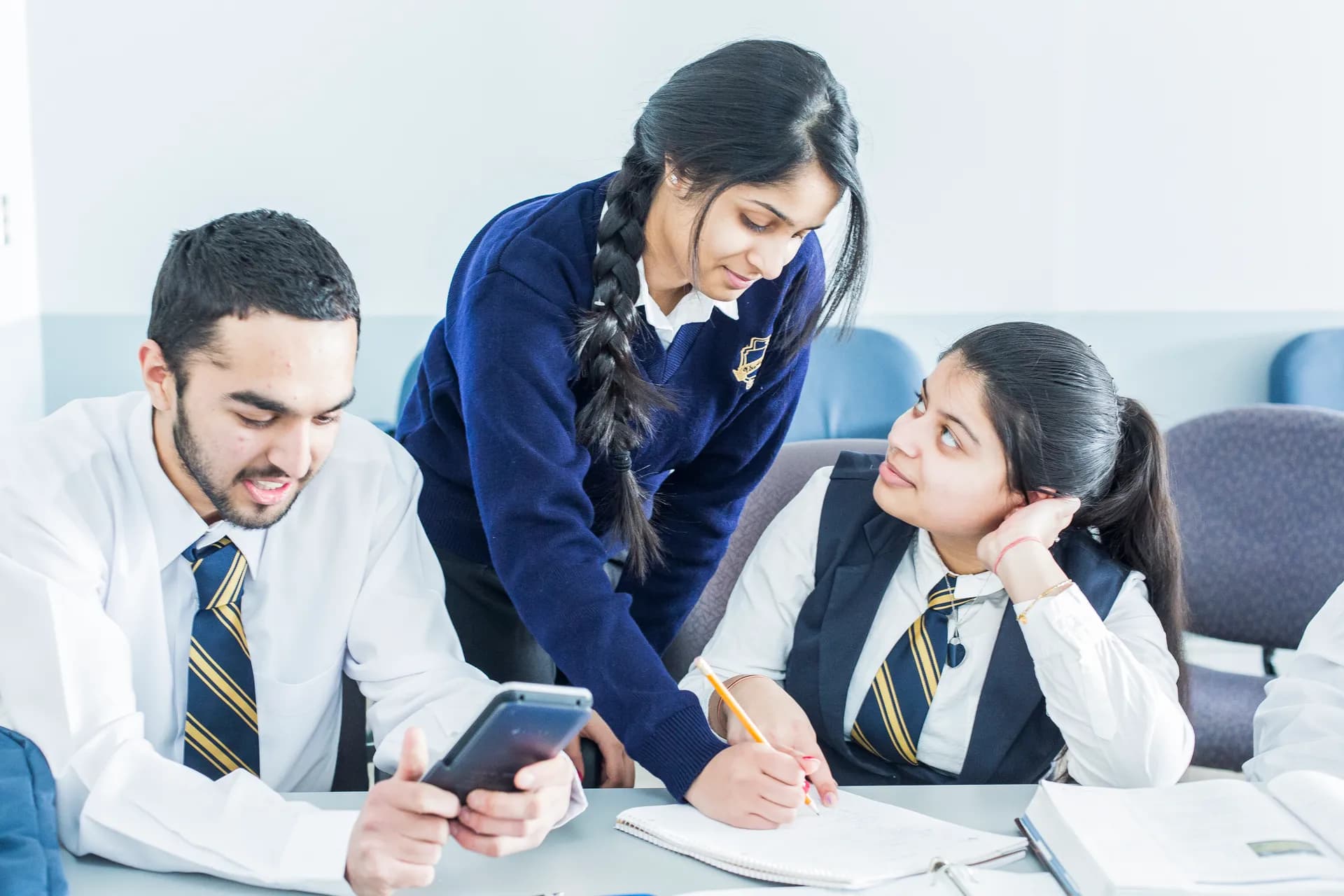 Three Vaughan College students in uniform work through a problem at a classroom table, one standing to explain