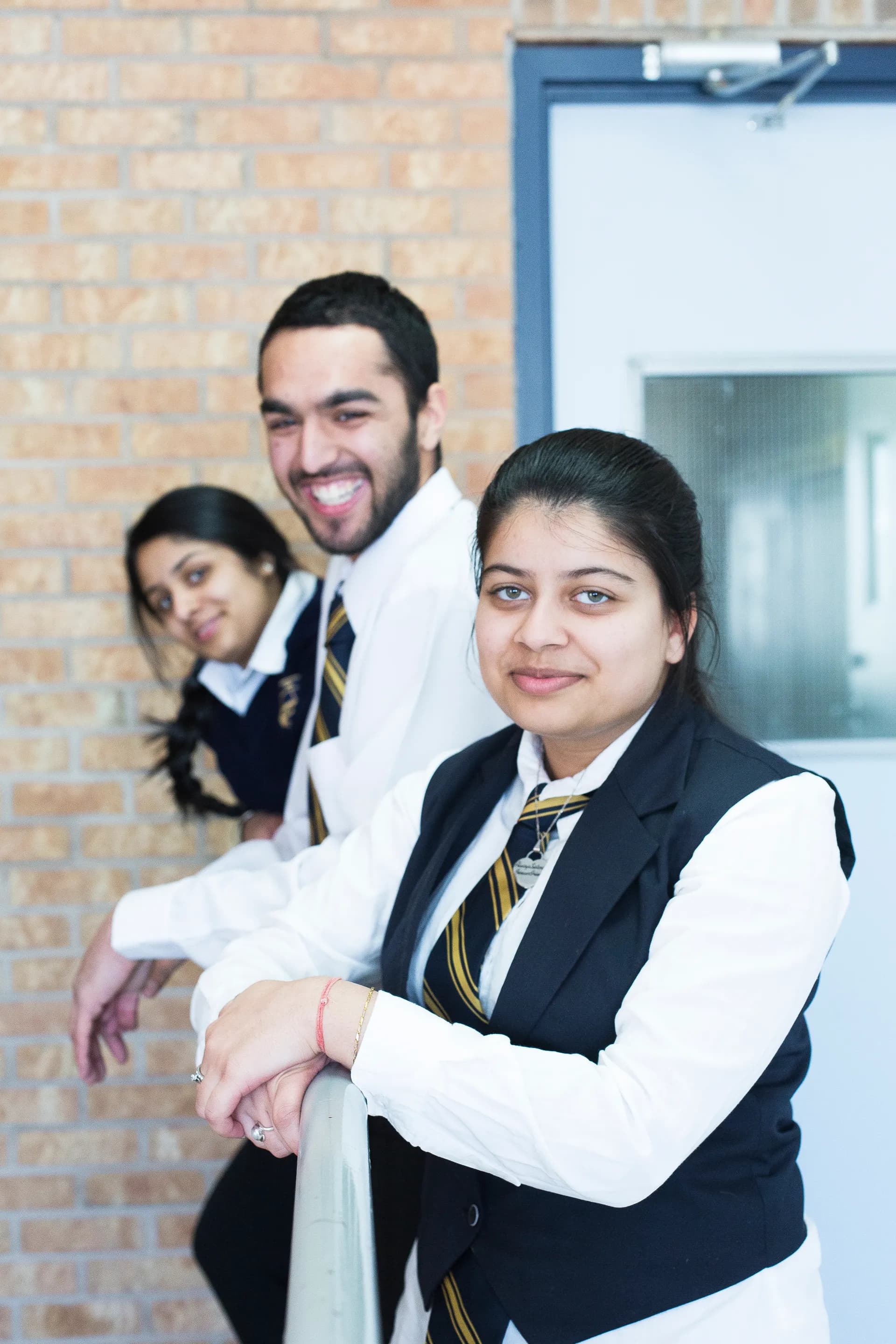 Three Vaughan College students in uniform smiling at the camera, leaning against a railing at school
